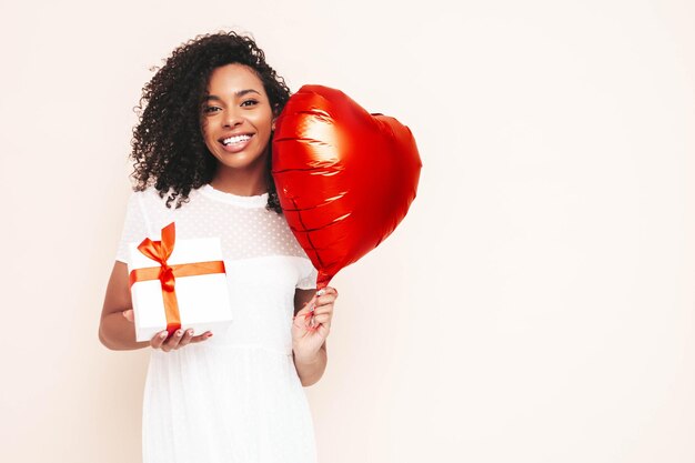 Mooie zwarte vrouw met afro krullen kapsel glimlachend model gekleed in witte zomerjurk Sexy zorgeloos vrouw poseren in de buurt van muur in studio Holding hart luchtballon en geschenkdoos geïsoleerd