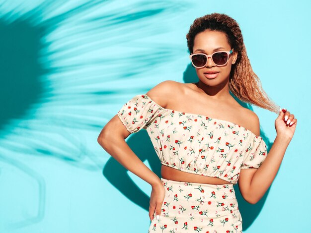 Mooie zwarte vrouw met afro krullen kapsel glimlachend model gekleed in zomer hipster kleding Sexy zorgeloos vrouw poseren in de buurt van blauwe muur in studio gelooid en vrolijk In zonnebril