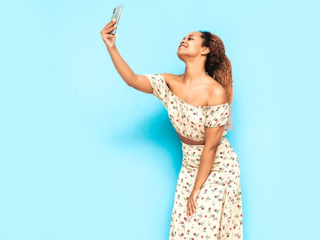 Mooie zwarte vrouw met afro krullen kapsel glimlachend model gekleed in zomerjurk sexy zorgeloze vrouw poseren in de buurt van blauwe muur in studio gelooid en vrolijk nemen selfie