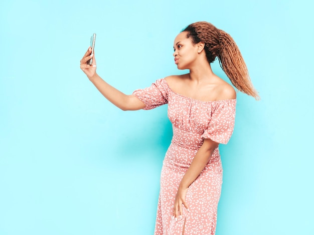 Mooie zwarte vrouw met afro krullen kapsel glimlachend model gekleed in zomerjurk Sexy zorgeloze vrouw poseren in de buurt van blauwe muur in studio gelooid en vrolijk nemen selfie