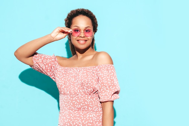 Mooie zwarte vrouw met afro krullen kapsel glimlachend model gekleed in zomerjurk Sexy zorgeloze vrouw poseren in de buurt van blauwe muur in studio gelooid en vrolijk