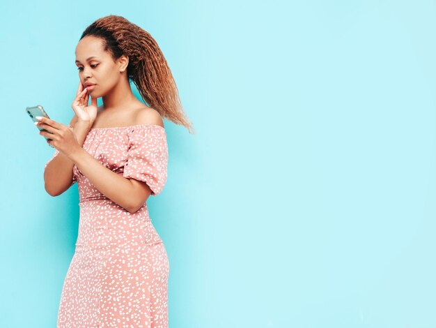 Mooie zwarte vrouw met afro krullen kapselGlimlachend model in trendy zomerkleren Sexy zorgeloze vrouw poseren in de buurt van blauwe muur in studio Smartphone-apps gebruiken kijken naar mobiel scherm