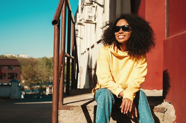 Mooie zwarte vrouw met afro krullen kapselLachende hipster model in gele hoodie Sexy zorgeloze vrouw poseren op de straat achtergrond in zonnebril vrolijk en gelukkig