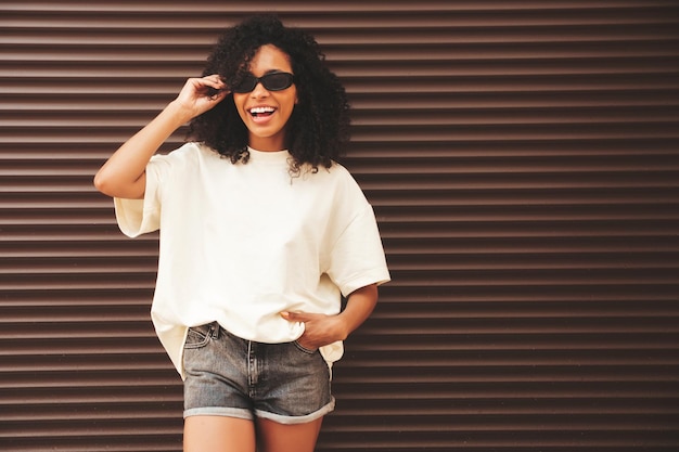 Mooie zwarte vrouw met afro krullen kapsellachende hipster model in witte tshirt sexy zorgeloze vrouw poseren in de straat in de buurt van bruine muur vrolijk en gelukkig in zonnebril