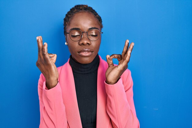 Mooie zwarte vrouw staande over blauwe achtergrond ontspannen en lachend met gesloten ogen doen meditatie gebaar met vingers yoga concept