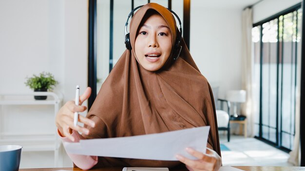 moslimdame draagt een koptelefoon met behulp van een computerlaptop praat met collega's over een verkooprapport in een videogesprek terwijl ze op afstand vanuit huis in de woonkamer werkt.