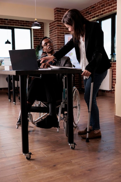 Multi-etnisch team van zakenmensen met een chronische beperking die samen aan een opstartproject werken. Vrouw met krukken en man in rolstoel brainstormen ideeën in gehandicaptenvriendelijk kantoor.