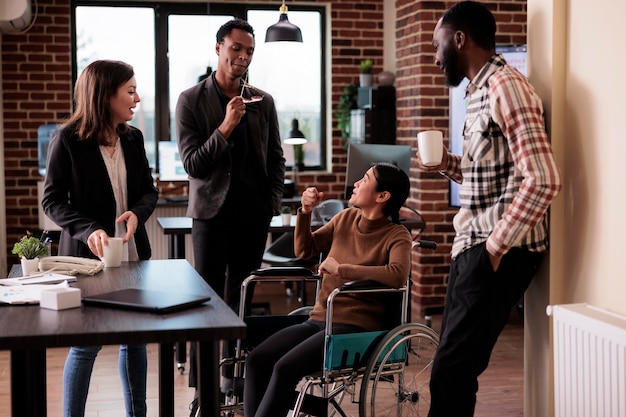 Multi-etnische groep collega's die lachen in het kantoor met een verlamde vrouw die in een rolstoel zit. Aziatische werknemer met chronische handicap en beperking die plezier heeft met collega's.