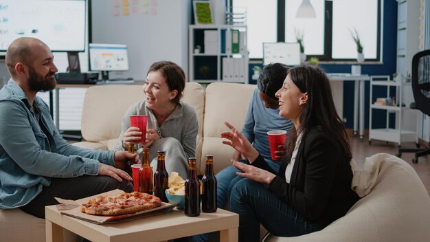 Multi-etnische groep collega's die plezier hebben na het werk op kantoor. Werkgenoten genieten na uren van feest met flessen bier, drankjes, pizza en snacks. Collega's lachen