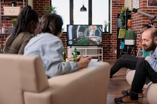 Multi-etnische groep vrienden die plezier hebben met shooter-competitie, thuis videogames spelen op tv-console met vrienden. genieten van gameplay bij het verzamelen, bier drinken.