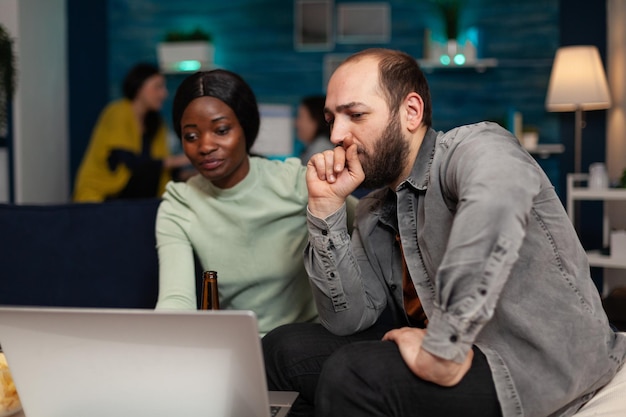 Multi-etnische vrienden zittend op de bank kijken naar online video op laptop en genieten van tijd samen doorbrengen tijdens het weekendfeest. Groep mensen die rondhangen, ontspannen in de woonkamer. vriendschap concept