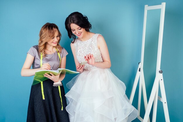 Naaister bruiloft consultant en bruid bespreken de details van trouwjurk in de studio op een blauwe achtergrond. kleermaker en klantbal in de kleedkamer naast de spiegel