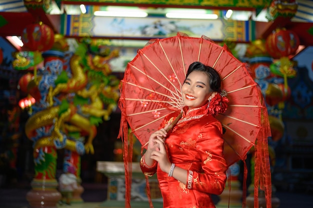 Nachtscène, portret aziatische mooie vrouw die een cheongsam draagt die lacht en poseert met papieren rode paraplu bij heiligdom op chinees nieuwjaar