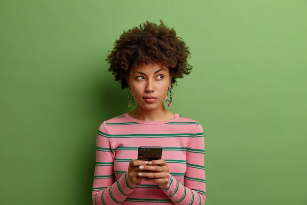 Nadenkend Afro-Amerikaanse vrouw gebruikt dating-app op smartphone wegkijkt draagt casual trui geïsoleerd over groene muur