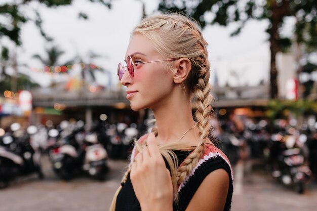 Nadenkend blonde vrouw in zwarte kleding poseren op straat achtergrond wazig. gebruinde dame met vlechten draagt roze zonnebril.