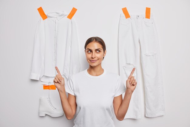 Nadenkend donkerharige mooie vrouw wijst wijsvingers hierboven adverteert kleding geïsoleerd op een witte achtergrond met gepleisterde t-shirt broek en laars Zwart-wit schot Je zou dit moeten dragen