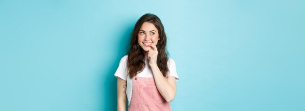 Nadenkend dwaas meisje met schattig krullend kapsel dat in de vinger bijt en naar links kijkt naar het promo-logo glimlachend