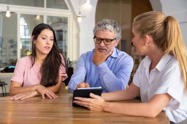 Nadenkend ernstige jonge vrouw en volwassen man ontmoeting met vrouwelijke professional, kijken en bespreken van inhoud op tablet