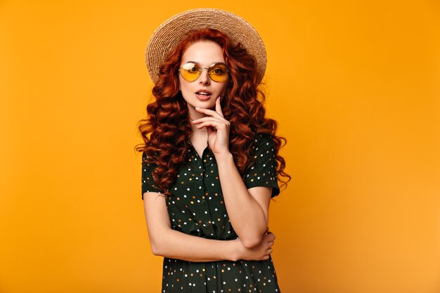 Nadenkend gember meisje poseren in zonnebril en strooien hoed. Vooraanzicht van romantische Europese dame geïsoleerd op gele achtergrond.