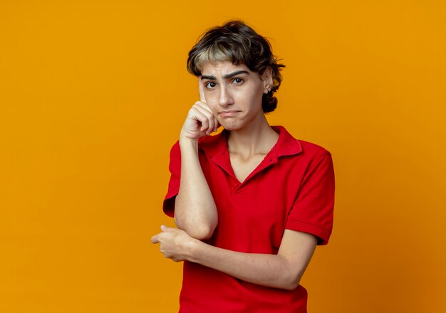 Nadenkend jong Kaukasisch meisje met pixiekapsel die vinger op tempel en hand onder elleboog zetten die op oranje achtergrond met exemplaarruimte wordt geïsoleerd