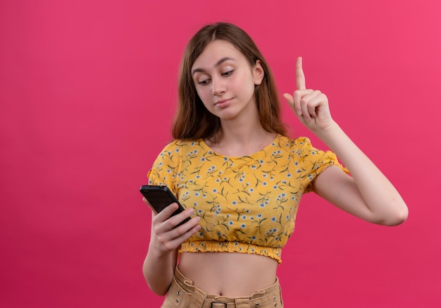 Nadenkend jong meisje dat mobiele telefoon houdt en vinger op geïsoleerde roze muur met exemplaarruimte opheft