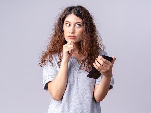 Nadenkend jong, mooi kaukasisch meisje met een mobiele telefoon die de hand op de kin houdt en naar de zijkant kijkt