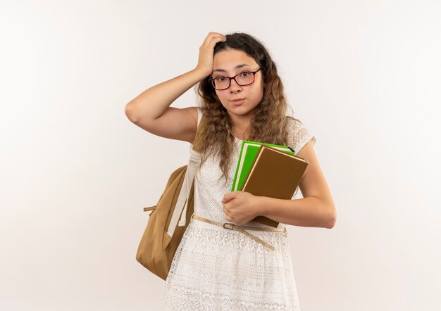 Nadenkend jong mooi schoolmeisje die glazen en rugtas dragen die boeken zetten die hand op hoofd zetten dat op witte muur wordt geïsoleerd