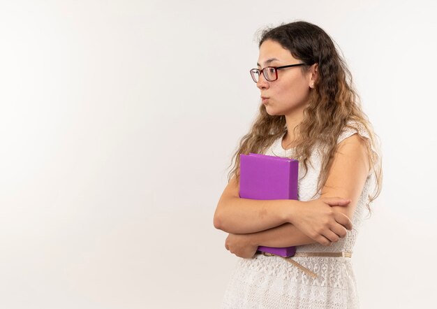 Nadenkend jong mooi schoolmeisje die glazen en rugtas dragen die zich in profielmening houden die boeken bekijken die kant bekijken die op witte muur wordt geïsoleerd