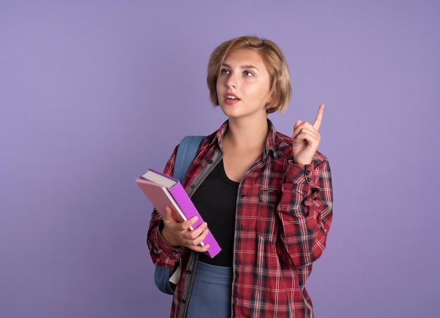 Nadenkend jong slavisch studentenmeisje dat een rugzak draagt, houdt een boek en een notitieboekje omhoog kijkend naar de zijkant