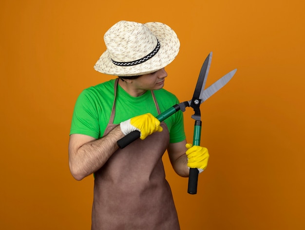 Gratis foto nadenkend jonge mannelijke tuinman in uniform dragen tuinieren hoed met handschoenen houden en kijken naar tondeuse geïsoleerd op oranje