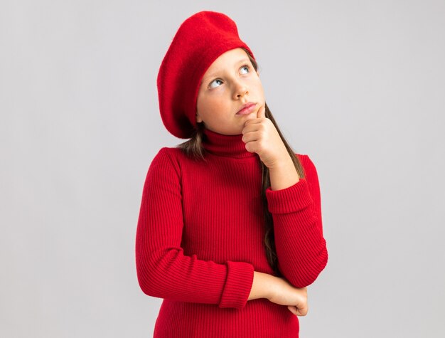 Nadenkend klein blond meisje met een rode baret die omhoog kijkt en de hand op de kin houdt, geïsoleerd op een witte muur met kopieerruimte