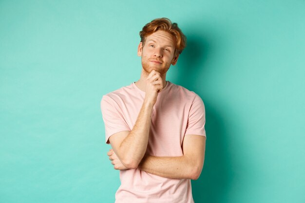 Nadenkend knappe man met rood haar en baard kijken naar de linkerbovenhoek, keuze maken en nadenkend kijken, staande in t-shirt op mint achtergrond.