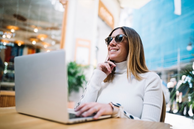 Nadenkend lachende jonge vrouw met behulp van laptop aan tafel in de straat café