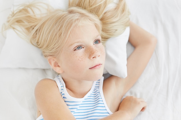 Nadenkend meisje met blauwe ogen en lange wimpers, met lang blond haar, zeeman T-shirt dragen, liggend op een wit kussen, opzij op zoek