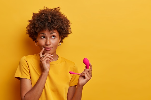 Nadenkend model met donkere huid heeft waterdichte g-spot vibrator op app-bediening op afstand voor stimulatie tijdens vrijen of solo spelen, verbonden met mobiele telefoon via bluetooth, lege ruimte op geel