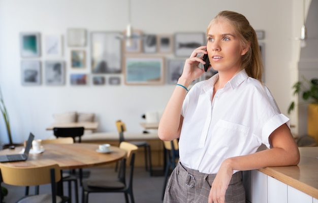 Nadenkend mooie jonge vrouw, gekleed in een wit overhemd, praten over de mobiele telefoon, permanent in co-werkruimte en wegkijken