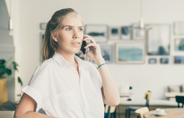 Nadenkend mooie jonge vrouw, gekleed in een wit overhemd, praten over de mobiele telefoon, permanent in co-werkruimte en wegkijken