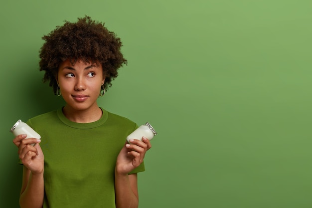 Nadenkend mooie vrouw geeft om gezondheid, houdt twee glazen flessen gezonde voedingsstof verse biologische yoghurt vast, geniet van het eten van zuivelproducten, draagt een groen t-shirt, vormt binnen, kopieert ruimte opzij voor promo