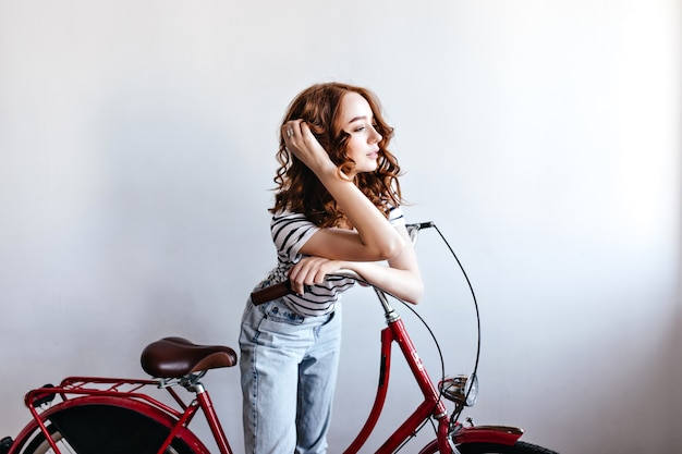 Nadenkend roodharige vrouwelijke model poseren met fiets. binnen schot van aantrekkelijke krullende vrouw naast de fiets.