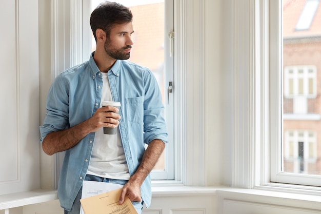 Nadenkend voorzitter met donkere haren, gekleed in een stijlvol overhemd, houdt koffie voor afhaalmaaltijden, papieren, studeert belastingen