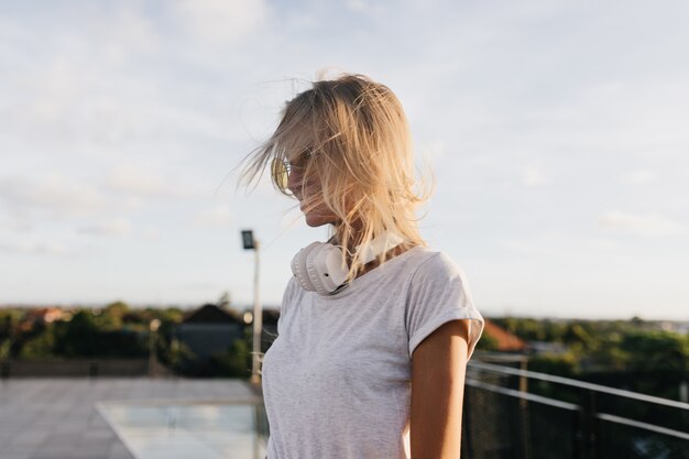 Nadenkend vrouw in wit t-shirt wegkijken tijdens een wandeling door de stad in de avond. stijlvolle blonde meisje in koptelefoon poseren op hemelachtergrond.