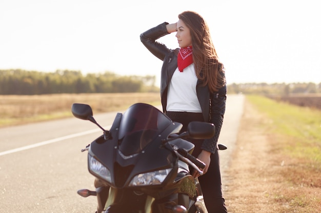 Nadenkend vrouwelijke bestuurder in stijlvolle kleding, poses op snelle motor, kijkt bedachtzaam opzij