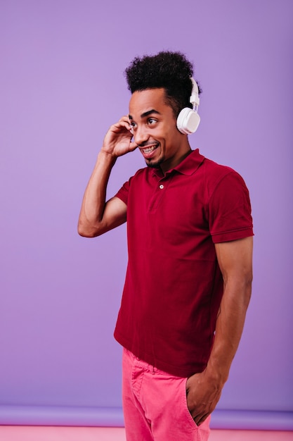 Nadenkende Afrikaanse man in rood t-shirt op zoek in de verte met een glimlach. Binnenfoto van zwart mannelijk model met grappig kapsel draagt een koptelefoon.