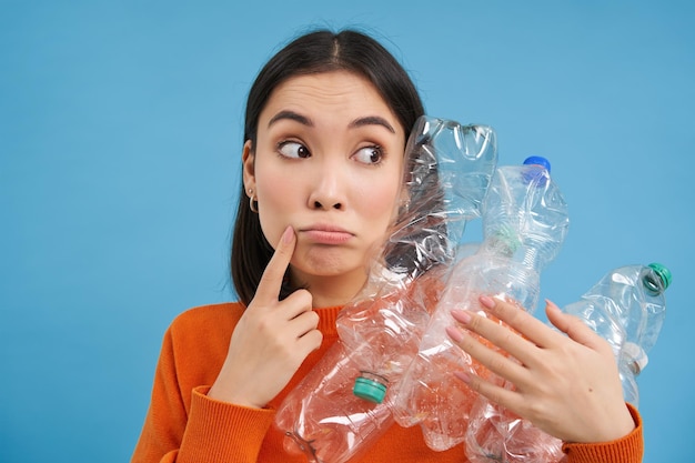 Gratis foto nadenkende aziatische vrouw die plastic flessen vasthoudt die opzij kijkt en nadenkt over het recyclen van sorteerga