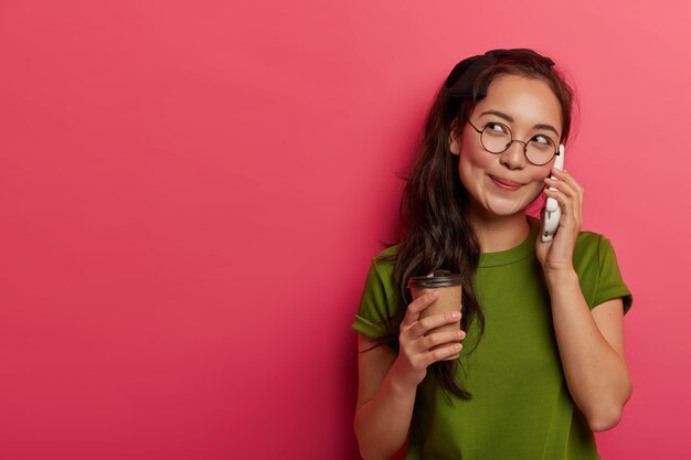 Nadenkende blije Aziatische tiener roept aangename herinneringen op tijdens telefoongesprekken, drinkt koffie en praat via mobiele telefoon, gefocust opzij, gekleed in vrijetijdskleding, voelt zich herbeleefd en ontspannen