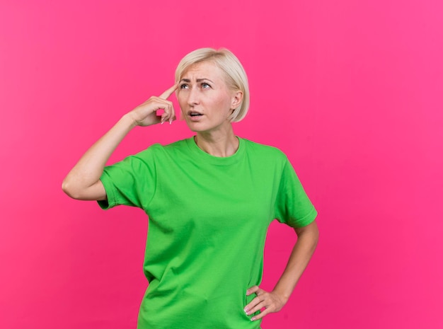 Nadenkende blonde Slavische vrouw van middelbare leeftijd die hand op taille houdt die omhoog doet denken gebaar dat op roze muur met exemplaarruimte wordt geïsoleerd