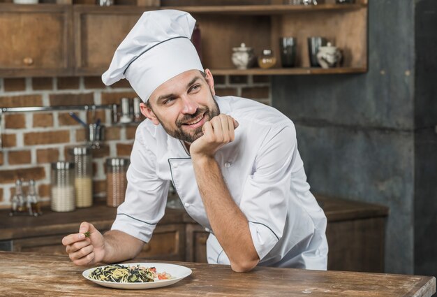 Nadenkende glimlachende mannelijke chef-kok die op de teller met een schotel in de keuken leunt