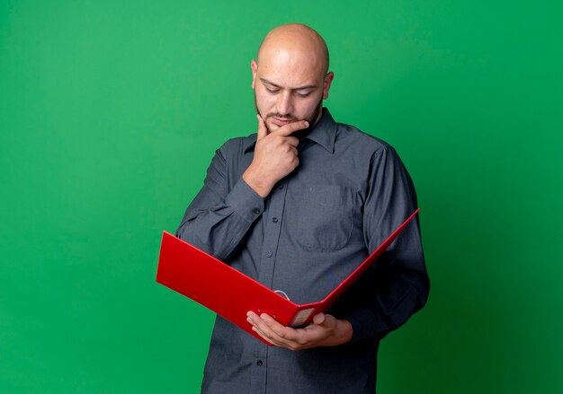 Nadenkende jonge kale callcentermens die hand op kin houdt en open map bekijkt die op groene muur wordt geïsoleerd