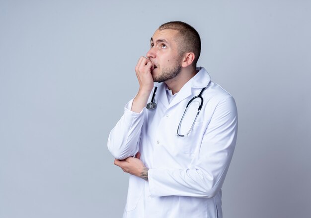 Nadenkende jonge mannelijke arts die medisch kleed en een stethoscoop om zijn hals draagt die zijn vingers bijt en omhoog geïsoleerd op witte muur kijkt