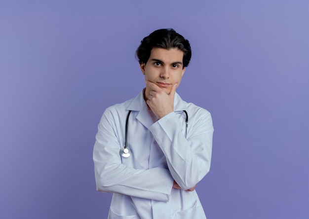 Nadenkende jonge mannelijke arts die medische mantel en stethoscoop draagt die kin raakt die op purpere muur met exemplaarruimte wordt geïsoleerd